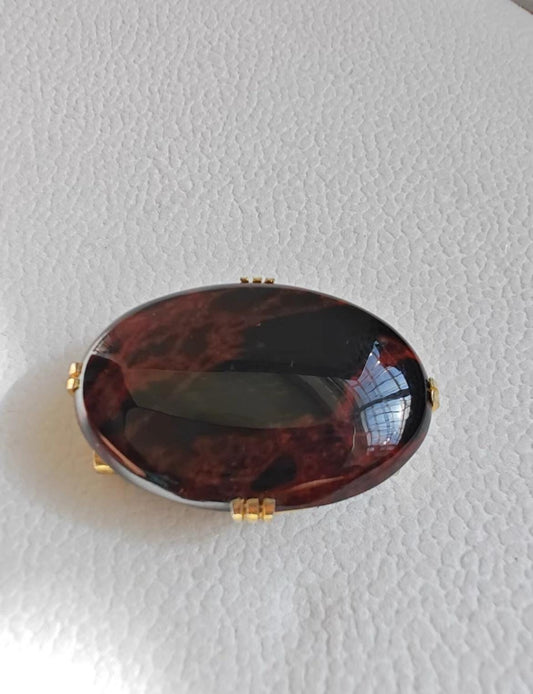 May include: An oval-shaped brooch with a dark red and black marbled design. The brooch is framed with gold-colored metal accents. The surface of the brooch reflects light, and the overall design is elegant.