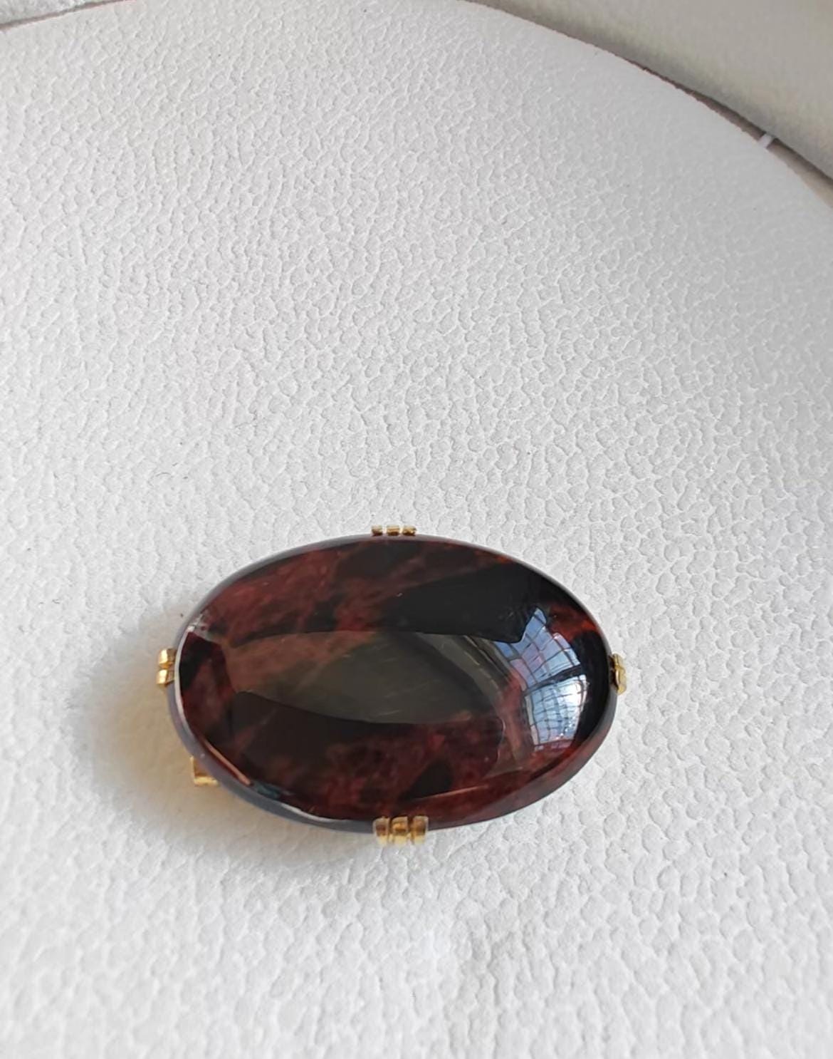 May include: Oval-shaped brooch with a dark red and brown stone, set in a gold-colored metal frame. The stone has a glossy finish, reflecting light. The brooch is a piece of jewelry.