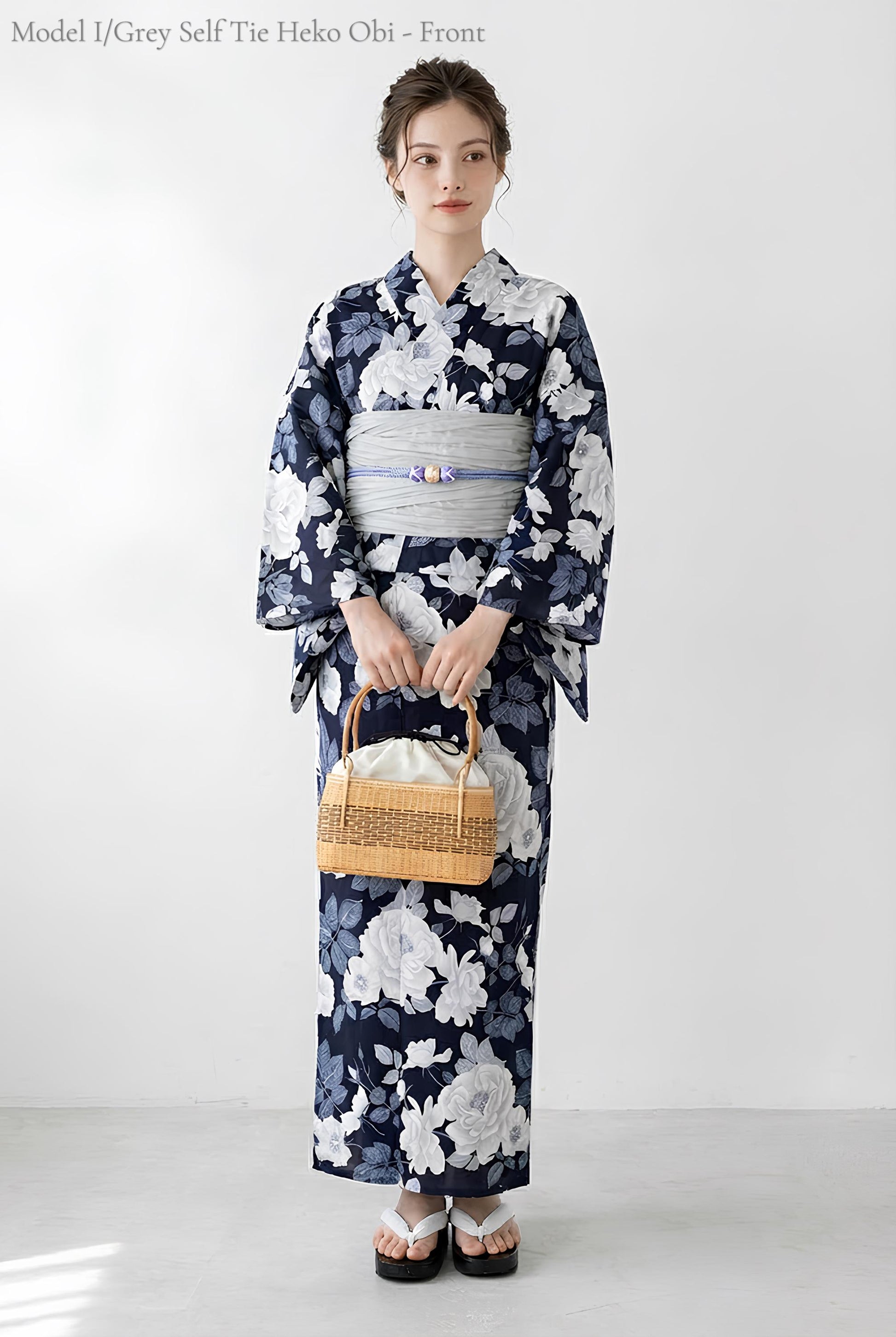 May include: A navy blue and white floral print yukata with a grey obi. The yukata features a large white floral pattern. A woven basket bag is held in front. The model is wearing white and black sandals. The text "Model I/Grey Self Tie Heko Obi - Front" is at the top.